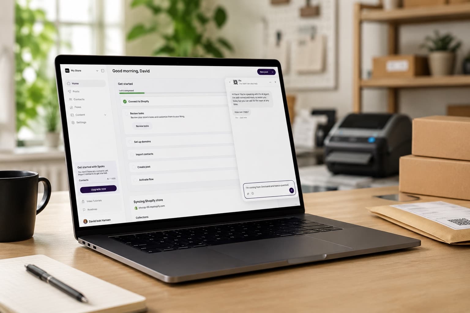 The Spoks dashboard open on a laptop at a Shopify packing desk, with a chat window where a merchant asks the Spoks team for help moving from Omnisend